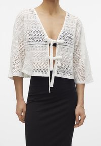 Wit kanten cropped top met wijde mouwen, voorzien van een diepe V-hals en striksluiting aan de voorkant. Gedetailleerde bloemige en geometrische patronen.