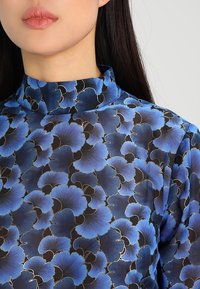 Halskrageblus med ett blått blommönster på en svart bakgrund, med en slät tygtextur och en subtil lyster.