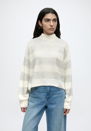 Mujer con un suéter de manga larga a rayas en crema y gris claro, con vaqueros de tiro alto azul claro, de pie frente a un fondo blanco liso.