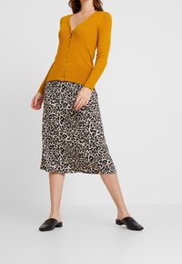 Senapsgul rippad cardigan med v-ringad hals och knappstängning, ihop med en beige kjol med svart leopardmönster och svarta slipskor.
