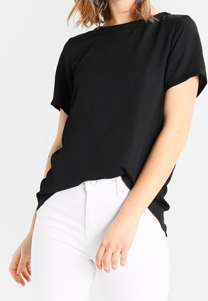 Kobieta ubrana w luźną czarną bluzkę z krótkim rękawem, lekko włożoną do białych spodni z wysokim stanem na jednolitym tle.