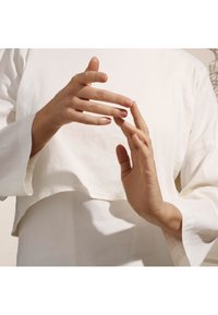 Weißes, leichtes Oberteil aus Stoff mit einem cropped Design und weiten Ärmeln. Die Hände gestikulieren, zeigen manikürte Nägel und eine natürliche Hauttextur.