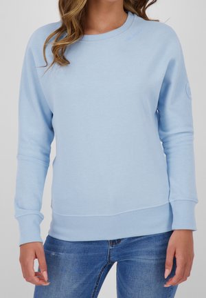 Frau trägt ein hellblaues Sweatshirt mit Rundhalsausschnitt, gerippten Bündchen und Saum, kombiniert mit blauen Jeans, steht vor einem schlichten Hintergrund.