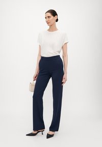 White ribbed top with short sleeves, paired with navy high-waisted trousers. Black pointed heels and a light beige handbag complete the outfit.