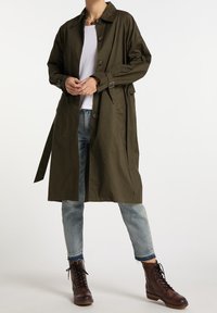 Płaszcz trencz w kolorze oliwkowej zieleni wykonany z lekkiego materiału, z kołnierzem, zapięciem na guziki i paskiem. Połączony z jasnoniebieskimi dżinsami i brązowymi butami.