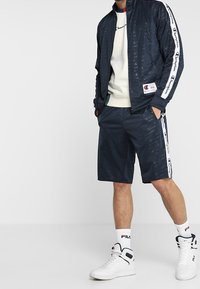 Marinblå idrottsjacka med dragkedja, mönstrad tyg och vit logotape; tillsammans med matchande shorts och vita sneakers med marinblå accenter.