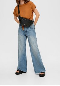 Svart läder axelremsväska med justerbar rem, bärs över en brun t-shirt; kombinerad med ljusblå vida jeans och svarta platåsandaler.