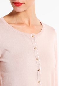 Ljusrosa kofta med långa ärmar och rund halsringning, försedd med knappslå framtill med gulddetaljer och en mjuk stickad struktur.