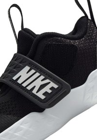 Baskets Nike noir et blanc avec une grande sangle sur le dessus affichant "NIKE" en lettres grasses et des languettes à boucle au talon et à la languette.