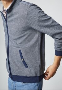 Chaqueta de cremallera en espiga azul marino y gris con puños y dobladillo acanalados, que cuenta con bolsillos laterales con acentos de botón. Tejido suave y texturizado.