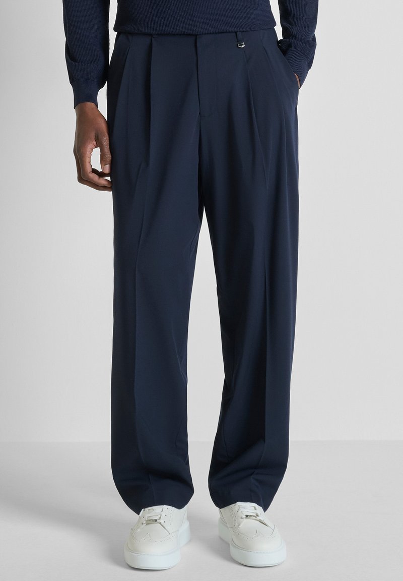 Hombre con pantalones anchos plisados de color azul marino y zapatillas blancas, con las manos en los bolsillos, de pie frente a un fondo liso.