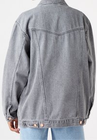 Grå denimjacka med en avslappnad passform, synlig sömnad och kopparfärgad hårdvarudetaljer vid ärmsluten och nederkant. Har en klassisk krage och bakstycke.