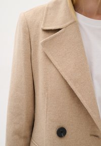 Bežs vilnas mētelis ar iedzimtu atloku, ar melnu pogu un gludu tekstūru. Valkāts virs baltas kreklas.