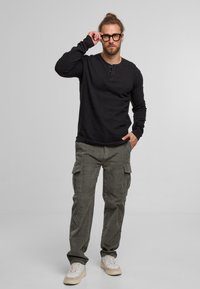 Schwarzes langärmeliges Henley-Shirt mit einer dreiknöpfigen Knopfleiste, kombiniert mit olivfarbenen Cargo-Hosen mit Seitentaschen und weißen Sneakers.