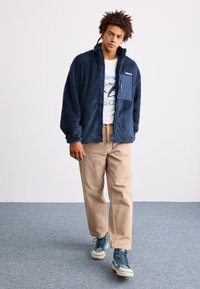 Mornarsko plava flis jakna s patentnim zatvaranjem, s džepom na prsima. Uparena s bež hlačama i plavim visokim tenisicama.