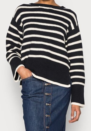 Pull en tricot noir et blanc à rayures avec une texture côtelée, des manches larges et un ourlet arrondi, associé à une jupe en denim avec des détails de boutons.