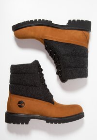 Bottes chevilles en daim marron avec une partie supérieure en feutre noir, une semelle en caoutchouc noir épaisse, un laçage à l'avant et un logo rond sur le côté.