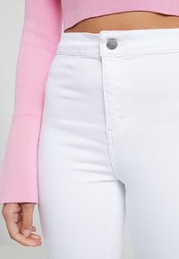 Bílé džíny s vysokým pasem a přiléhavým střihem, se zapínáním na stříbrný knoflík a prošitím kapes, doplněné růžovým topem s dlouhým rukávem.