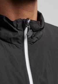 Chubasquero negro con un cuello alto, que presenta un cierre de cremallera blanca en contraste y un tirador de cremallera con forma de logo. Textura suave.