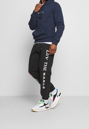 Felpa della marina con cordini, pantaloni tuta neri con la scritta "LUV THE W.O.R.L.D" e sneakers bianche con accenti colorati e suole testurizzate.