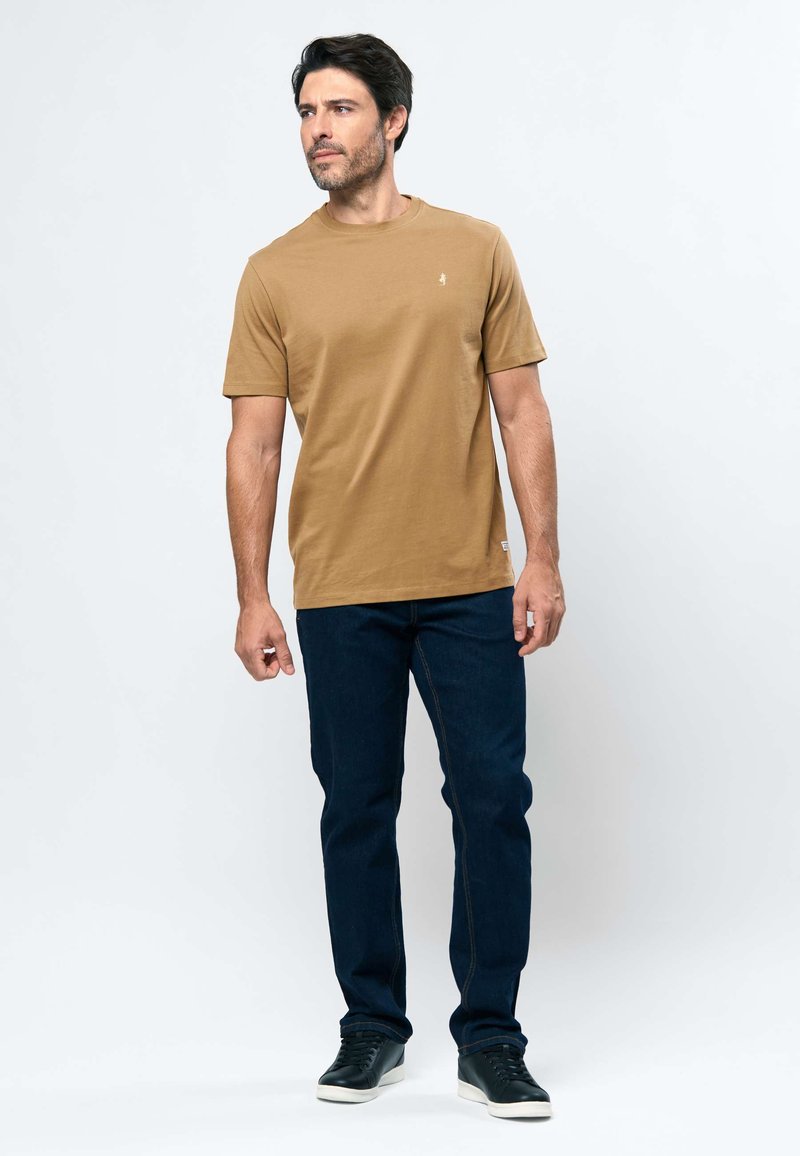 Homme debout, portant un t-shirt marron à manches courtes, un jean bleu foncé et des baskets noires, regardant vers sa gauche sur un fond blanc uni.