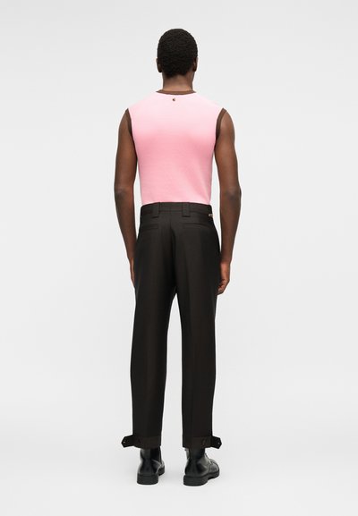 Άνδρας που φοράει ροζ αμάνικη μπλούζα με καφέ περιθώριο, μαύρο ραμμένο παντελόνι με γυρισμένα μπατζάκια και μαύρες δερμάτινες μπότες, στέκεται στραμμένος προς τα πίσω.