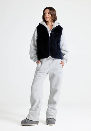 Vrouw draagt lichtgrijze hoodie en joggingbroek met zwarte fleece bodywarmer en grijze pantoffels, staat met handen in de zakken tegen witte achtergrond.