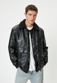 Chaqueta de cuero negra con un cuello texturizado negro, botones a presión, dos bolsillos delanteros y un diseño ajustado, combinada con jeans azul claro.