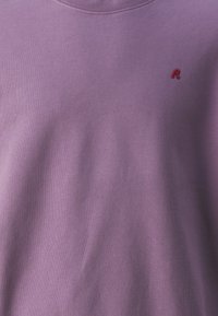Fioletowa bluza z miękkiego materiału, z okrągłym dekoltem i małym czerwonym logo "R" wyszytym po lewej stronie.