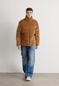Braune gepolsterte Jacke mit hohem Kragen, Reißverschluss und gestepptem Muster, getragen über abgetragenen blauen Jeans und weißen Schuhen.
