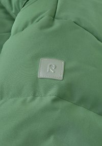 Zielony, otulający materiał z miękką fakturą. Zawiera kwadratową metkę wytłoczoną z wypukłym logo "R", uzupełnioną delikatnymi detalami przeszycia.