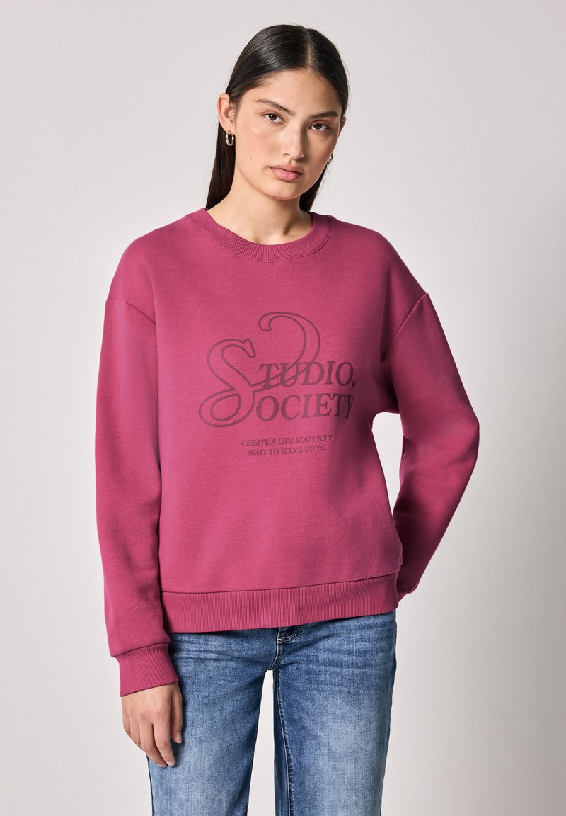 Felpa fucsia con scollo rotondo e spalle cadenti. Presenta un design grafico in nero con la scritta "STUDIO SOCIETY" e testo sottostante.