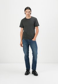 Graues Baumwoll-T-Shirt mit kurzen Ärmeln, kombiniert mit blauen Jeans. Das Modell trägt schwarze Schnürschuhe. Einfarbiger Hintergrund.