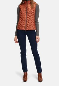 Kobieta w rdzawym, pikowanym kamizelce na szarym, prążkowanym, długim rękawie, ciemnoniebieskich dżinsach i brązowych botkach na białym tle.