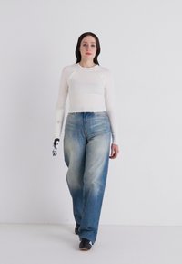 Weißes Langarmoberteil, tailliert mit glatter Textur. Hellblaue Jeans, lockere Passform mit einem verwitterten Design. Schwarze Schuhe mit weißen Akzenten.