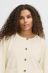 Camisa de botón color crema hecha de tejido texturizado, con botones de madera y grandes bolsillos frontales.