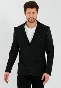 Zwarte zip-up jack met een textuur op de schouderpanelen, slim fit ontwerp en subtiele zakdetails, gedragen over een wit shirt.