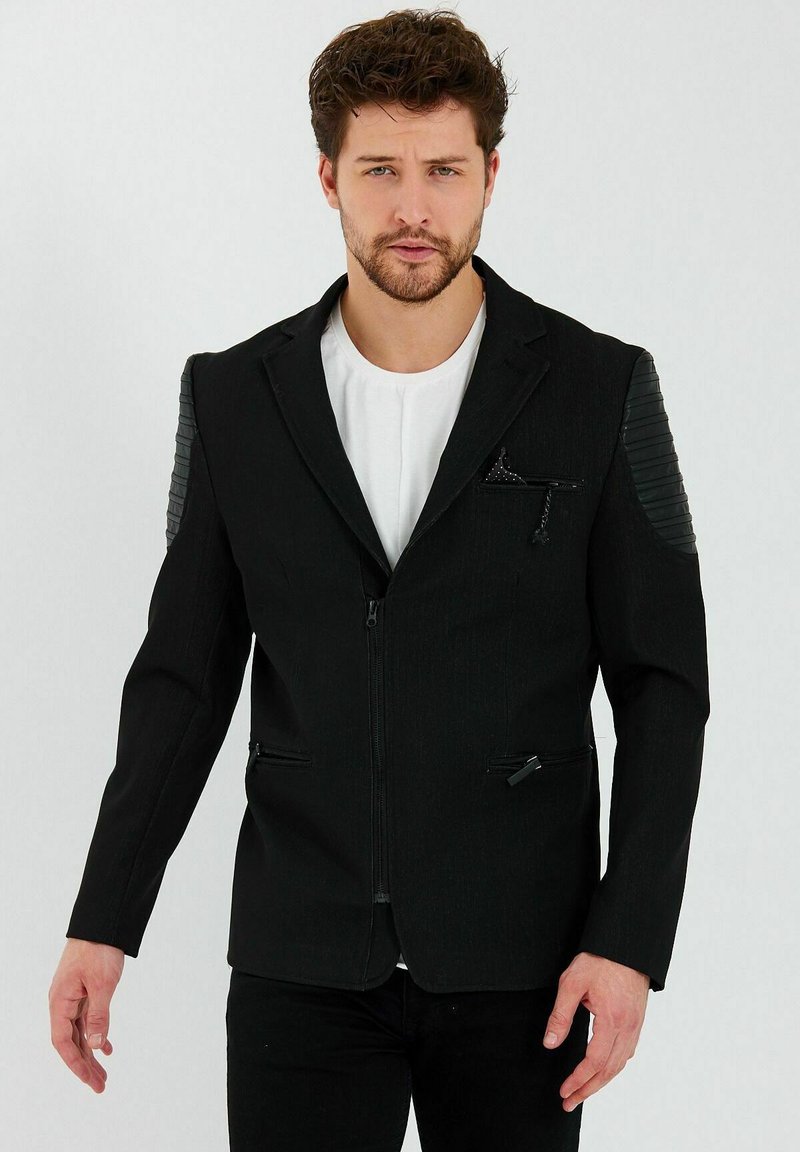 Zwarte zip-up jack met een textuur op de schouderpanelen, slim fit ontwerp en subtiele zakdetails, gedragen over een wit shirt.