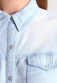 Ljusblå denimskjorta med spetsig krage, knappframsida och bröstfickor. Har silverfärgade tryckknappar och en slät textur.