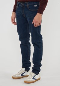 Jeans blu scuro con una vestibilità slim, chiusura a bottone e dettagli di cucitura visibili, abbinati a sneakers bianche e beige con accenti blu navy.