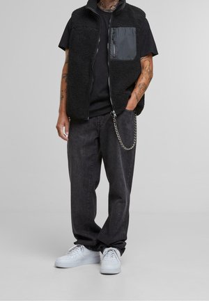 Personne portant un gilet polaire noir avec poche poitrine zippée, un t-shirt noir, un jean ample foncé avec une chaîne et des baskets blanches, debout devant un fond gris.