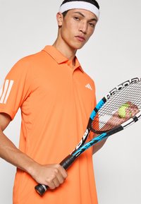 Polo de color naranja brillante con cuello y tres rayas blancas en la manga izquierda, sosteniendo una raqueta de tenis y una pelota, sobre un fondo neutro.