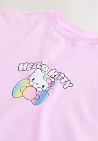 T-shirt rose clair avec un motif Hello Kitty tenant des formes aux couleurs pastel de l’arc-en-ciel et le texte « Hello Kitty » en lettres gonflées au-dessus.