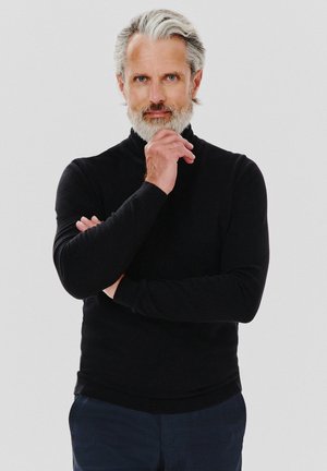 Czarny sweter z golfem wykonany z dzianiny, dopasowany krój, długie rękawy, noszony z ciemnymi spodniami w wzory; widoczne siwe włosy i broda.