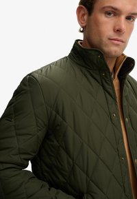 Hombre vistiendo una chaqueta acolchada verde oliva con cuello alzado sobre un suéter color beige con cremallera, contra un fondo blanco.