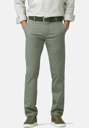 Man draagt een opgerolde lichtbeige overhemd, olijfgroen riem, saliegroene broek en olijfgroene sneakers met witte zolen, handen in de zakken.