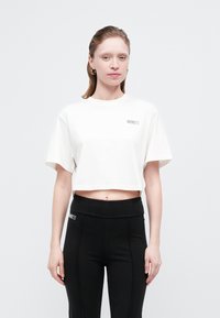 Femeie cu piele deschisă și păr brunet, îmbrăcată cu un tricou alb scurt și pantaloni negri cu talie înaltă, stând în fața unui fundal deschis simplu.
