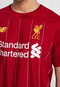 Röd Liverpool fotbollströja med vita vertikala ränder, broderad logotyp och sponsorlogotyper i vitt. Tillverkad av andningsbart material.
