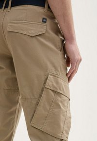 Beige cargo bukser med en struktureret stof, der har sidelommer og en baglomme. En mørk taljebånd komplementerer designet.