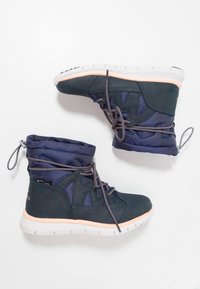 Botas de tobillo azul oscuro con parte superior de malla y cuero, cordones grises y una suela blanca y melocotón. Presenta un puño fruncido con un ajuste regulable.